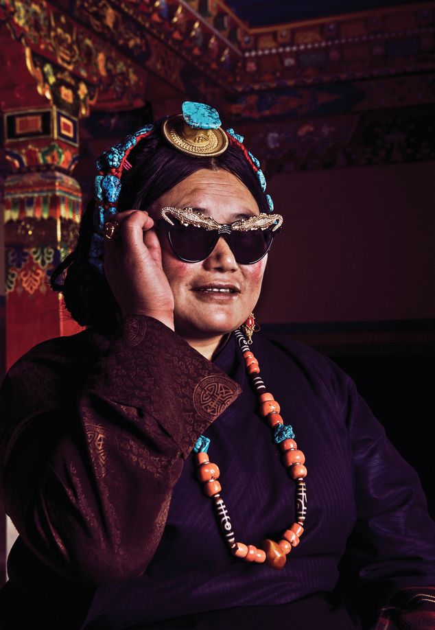 An elderly Tibetan woman in traditional outfit tries on a pair of glamorous sunglasses in “Modernizing the World Roof,” 2014