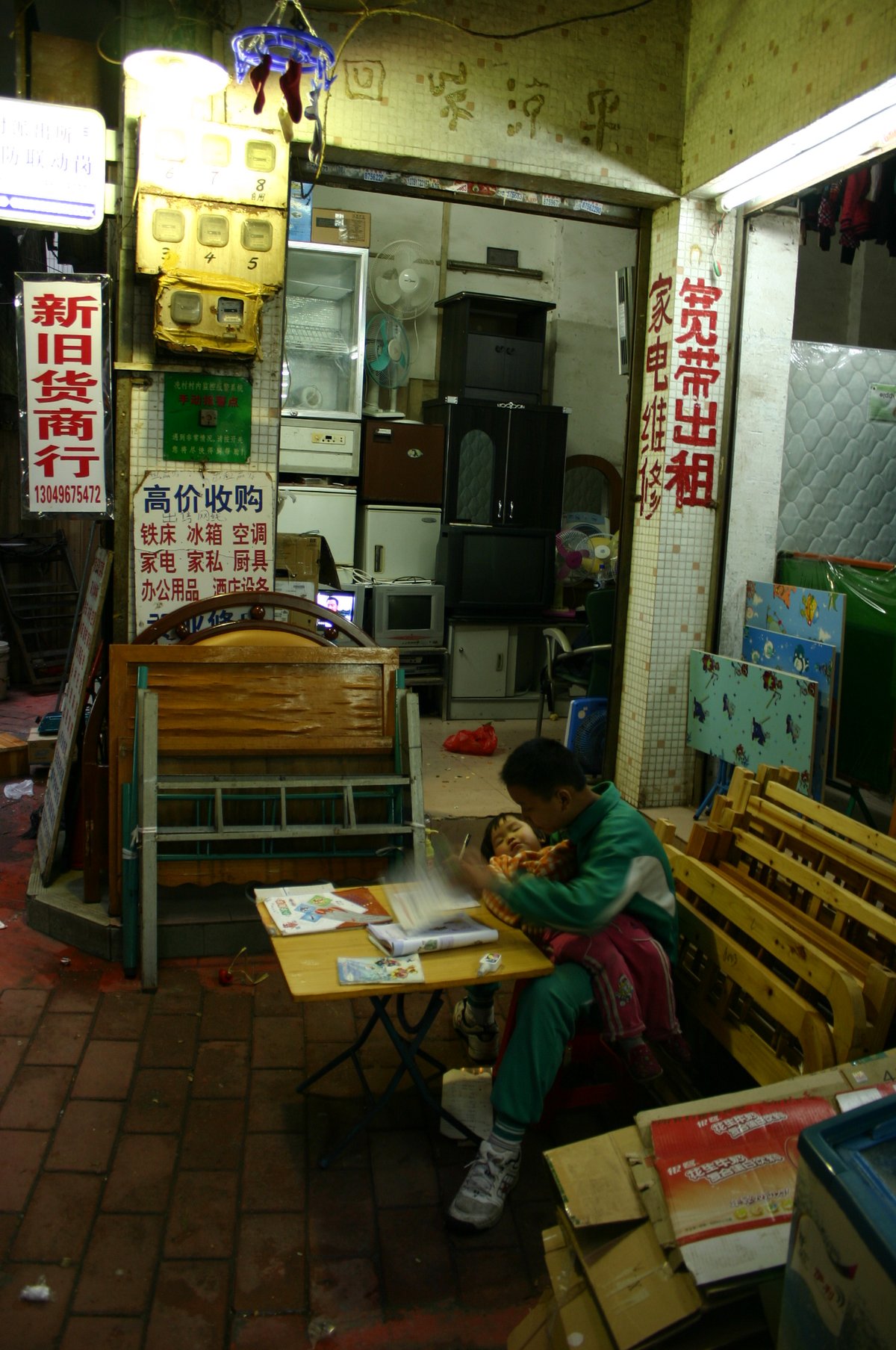 In Xiancun, a little boy holds his sleeping toddler sister in his arms. He completes his homework while waiting for their mother to finish work at the family food stall.