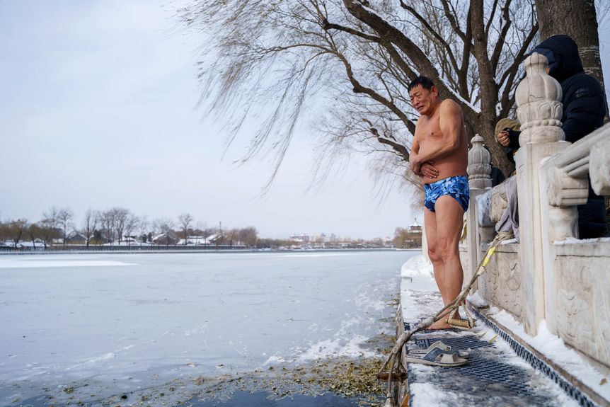A man in a blue swimsuit stands on a ledge by a frozen lake, First snow in Beijing in 2023