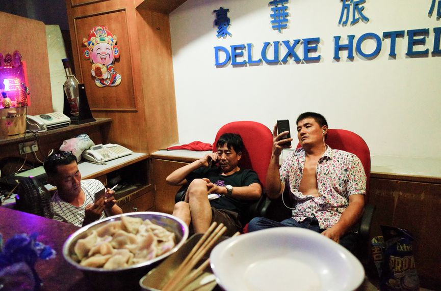 Two Chinese men sitting under a Deluxe Hotel sign with food in front, Yan Jiacheng "Deluxe Hotel"