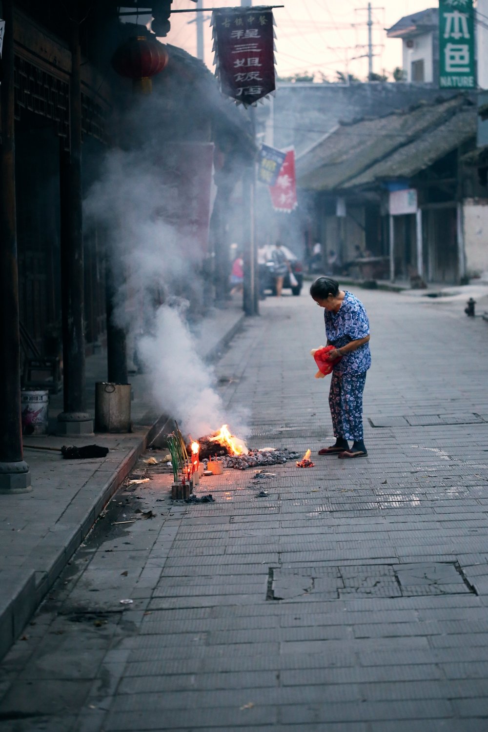 An elderly woman burning paper goblets for her deceased family members at sunset in Huaiyuan Town