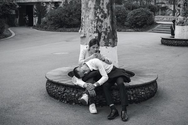 A Chines man taking a nap on a woman&#x27;s lap as she takes browses on her phone. 