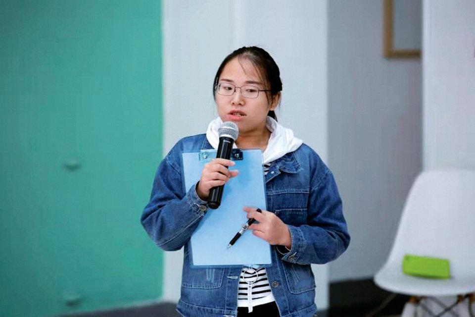 Tong Can holds up a microphone and stands up to speak at an event, while holding a clipboard and pen in her left hand.