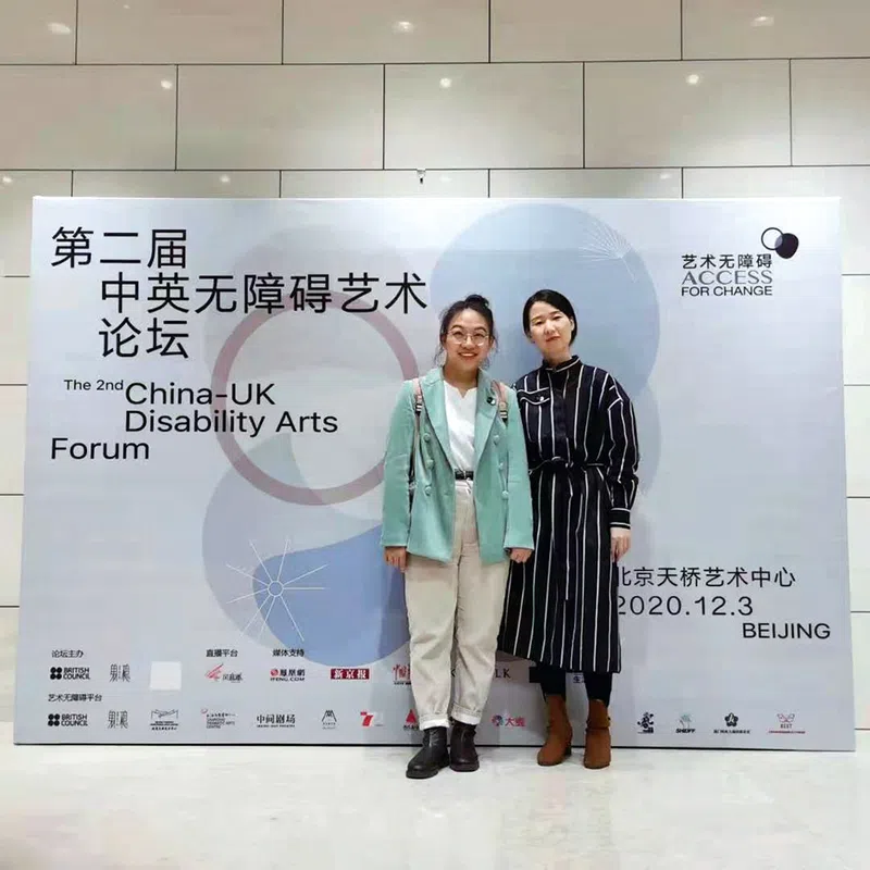 Peng Yujia and a woman take a photo in front of a Chinese-UK Disability Arts Forum banner.
