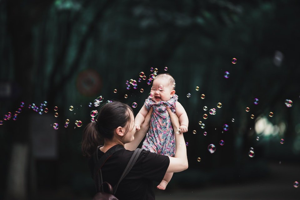 Mom holding baby in the air with bubbles floating around (Unsplash)